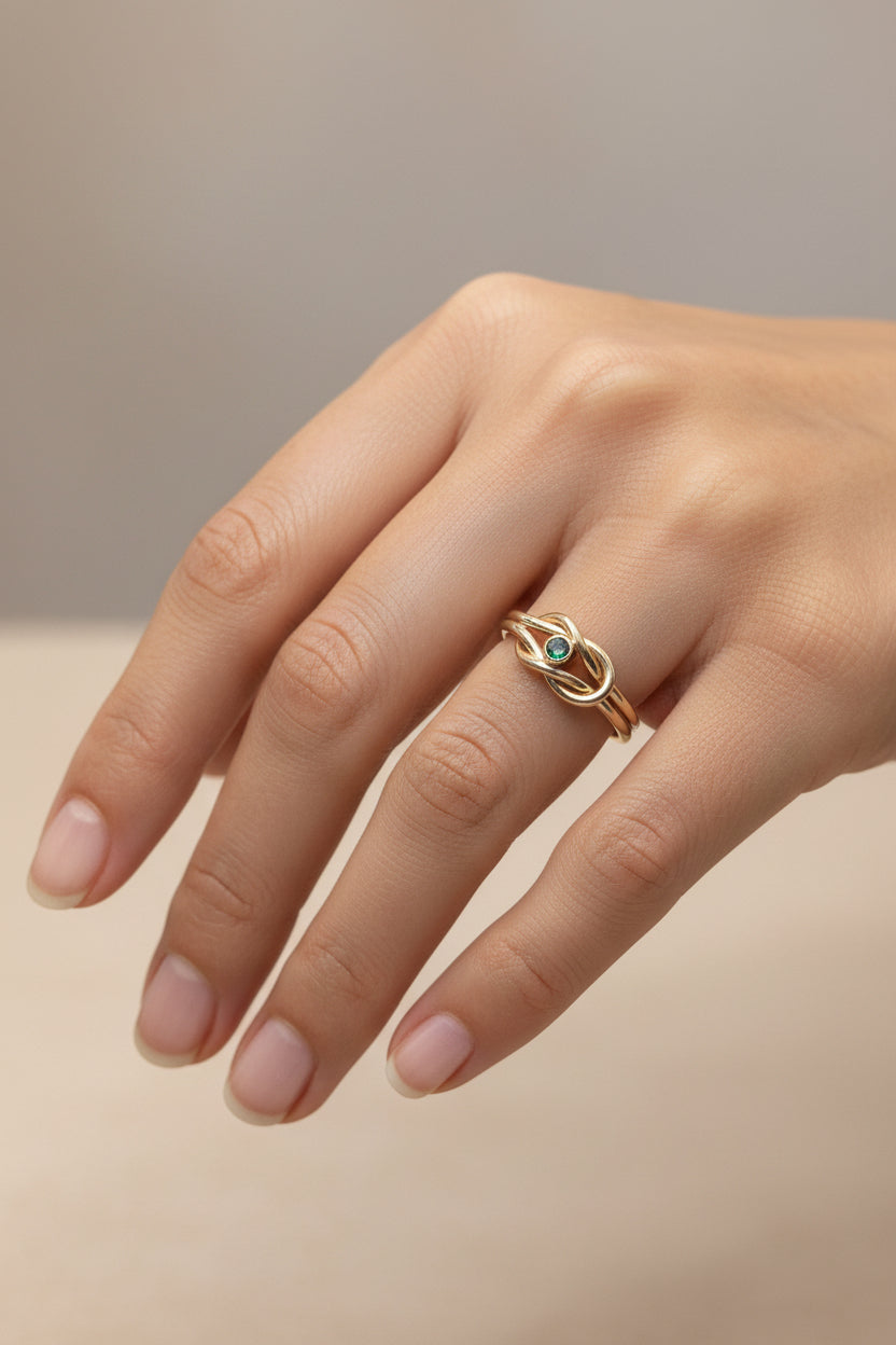 A close-up of a hand wearing a gold ring with a small green stone set in an intertwined band design, against a soft beige background.